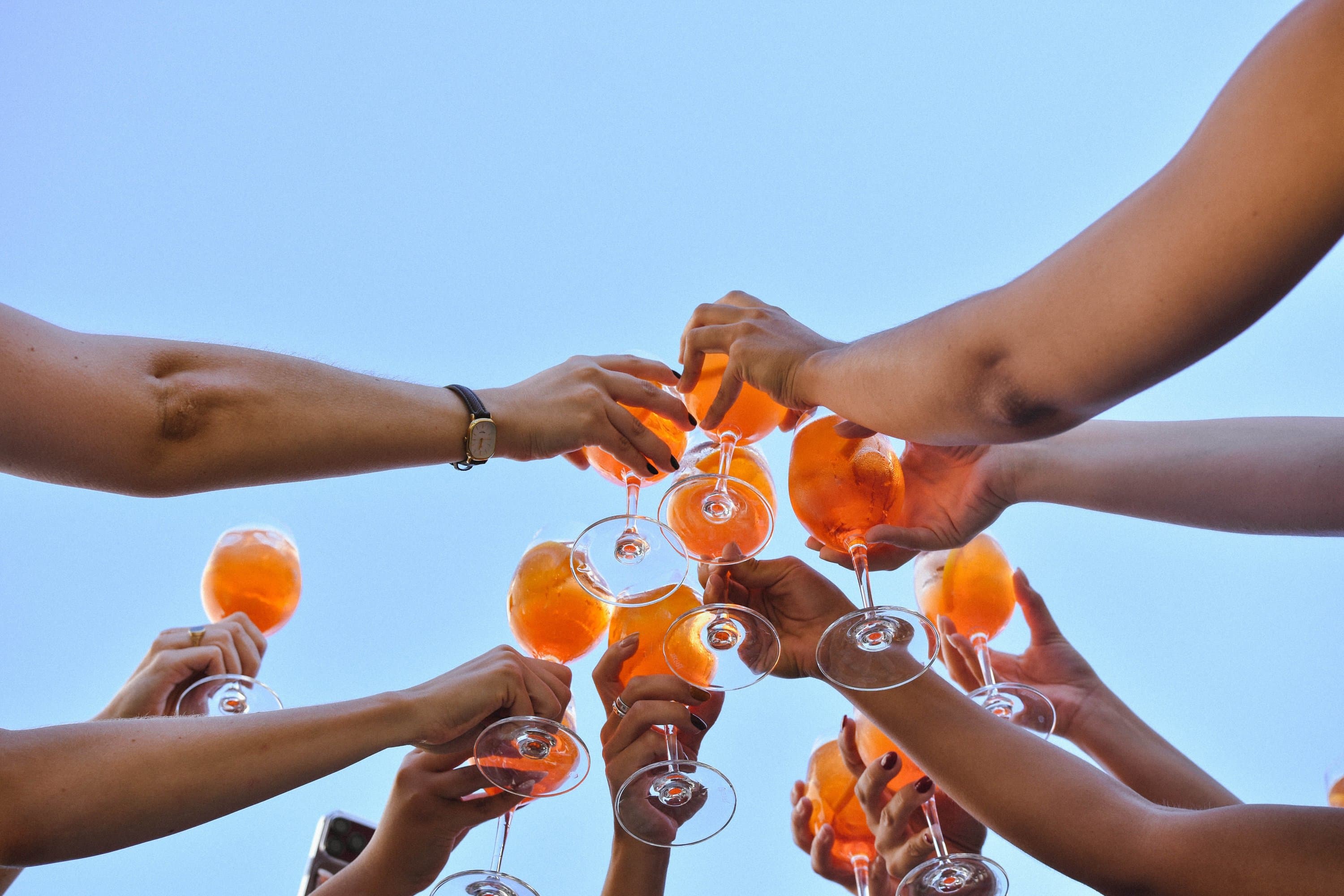 Hands toast with orange spritz glasses against a blue sky.